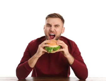 Young man eating tasty burger at table on white background