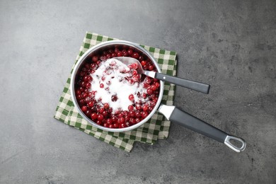 Photo of Making cranberry sauce. Fresh cranberries with sugar in saucepan and spoon on gray table, top view
