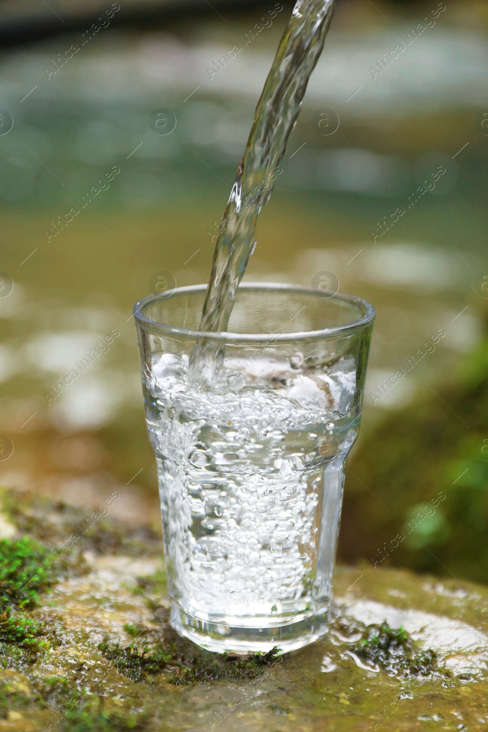 Photo of Fresh water pouring into glass on stone near river, closeup