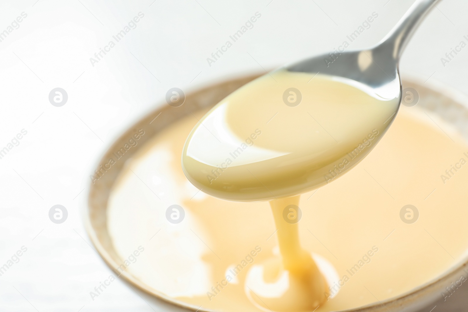 Photo of Spoon of pouring condensed milk over bowl on table, closeup with space for text. Dairy products