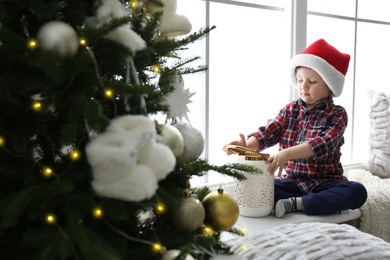Photo of Cute little boy in Santa hat playing with Christmas candleholder on window sill at home