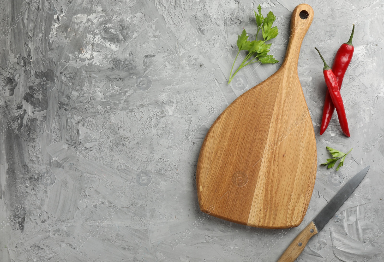 Photo of Cutting board, knife, chili peppers and parsley on grey textured table, flat lay. Space for text