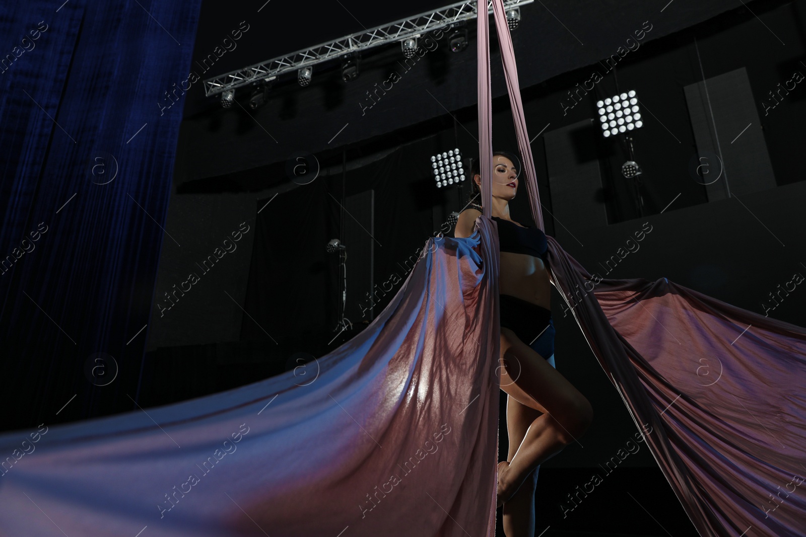 Photo of Young woman performing acrobatic element on aerial silk indoors, low angle view