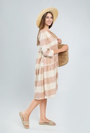 Young woman wearing stylish dress with straw bag on light grey background