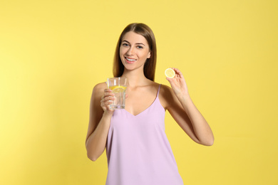 Young woman with glass of lemon water on yellow background