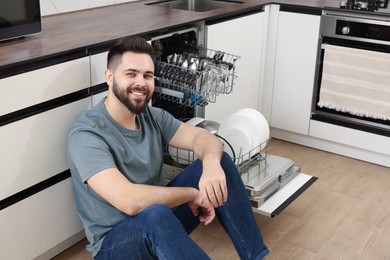 Smiling man sitting near open dishwasher in kitchen