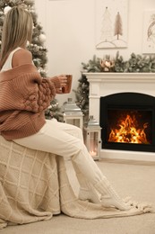 Photo of Woman with hot drink resting near fireplace in cozy room decorated for Christmas, back view