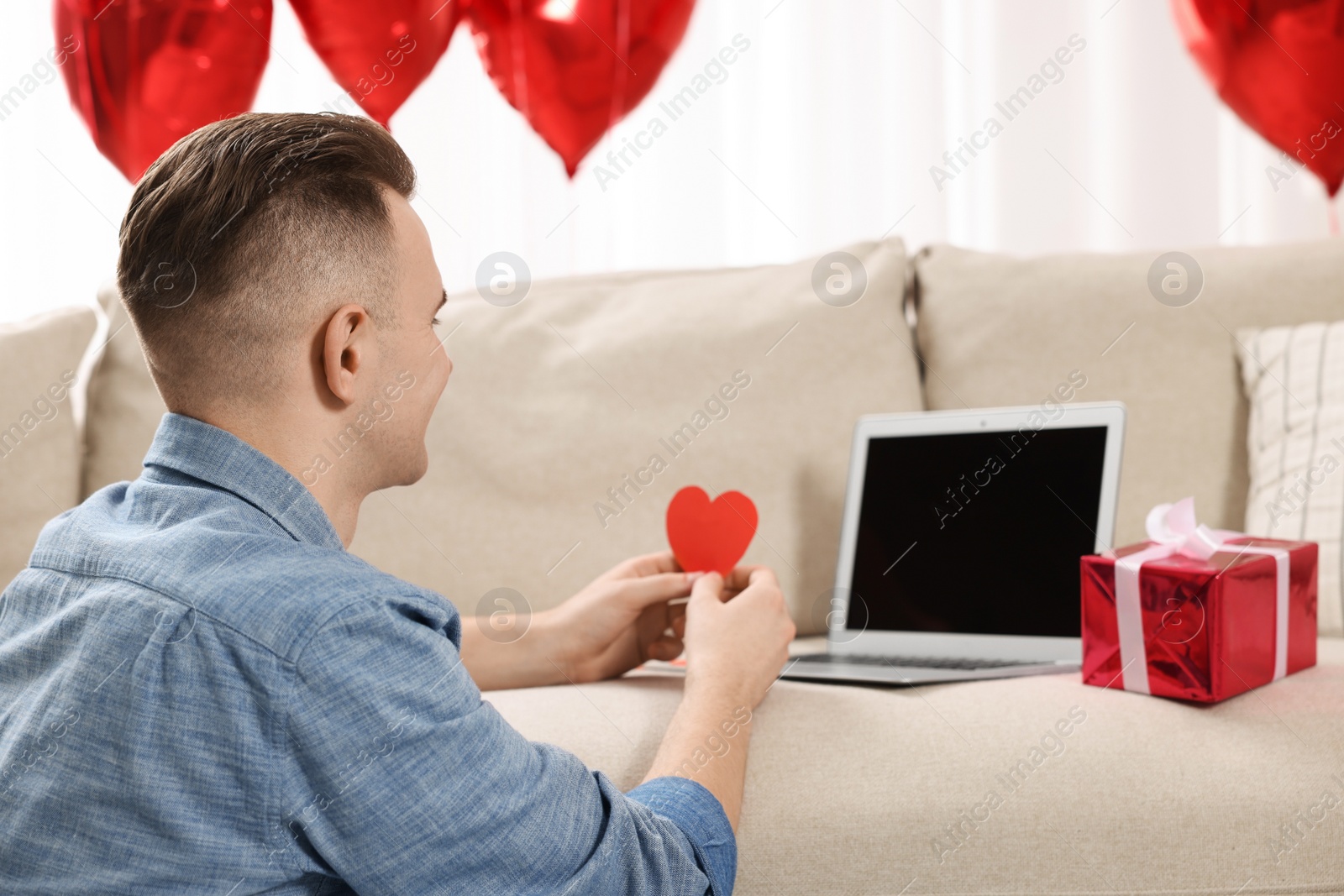 Photo of Valentine's day celebration in long distance relationship. Man holding red paper heart while having video chat with his girlfriend via laptop indoors