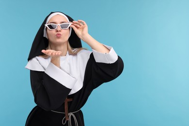 Woman in nun habit and sunglasses blowing kiss against light blue background. Space for text