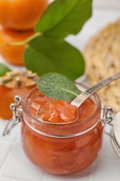 Jar and spoon of tasty persimmon jam on white tiled table, above view