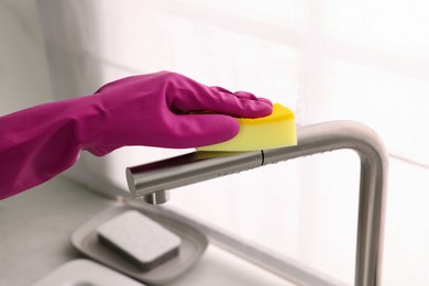 Photo of Woman in gloves cleaning faucet of kitchen sink with sponge, closeup