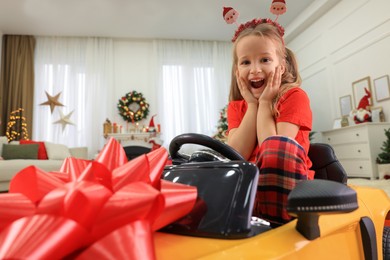 Photo of Excited little girl sitting inside toy car in room decorated for Christmas
