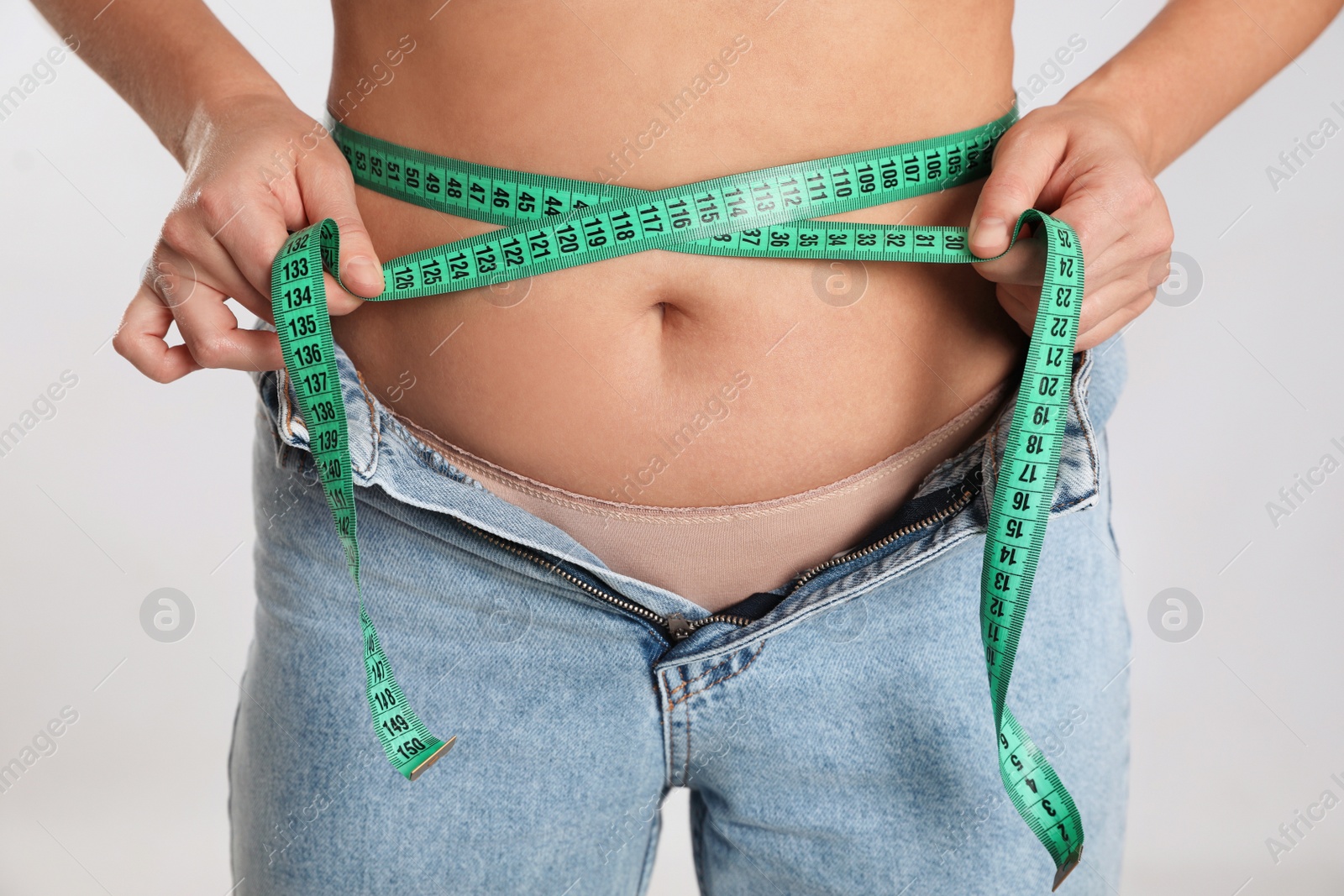 Photo of Woman in unfit jeans measuring her waist on light background, closeup. Weight loss concept