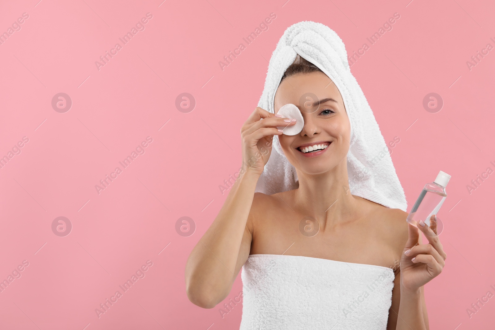Photo of Smiling woman removing makeup with cotton pad and holding bottle on pink background. Space for text