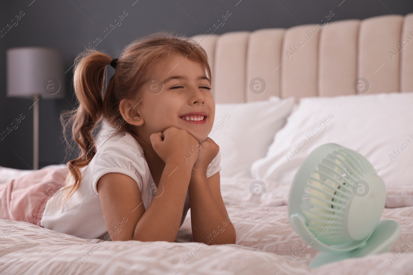 Photo of Little girl enjoying air flow from portable fan on bed in room. Summer heat