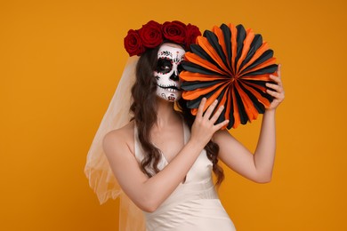 Photo of Young woman in scary bride costume with sugar skull makeup, flower crown and paper decoration on orange background. Halloween celebration