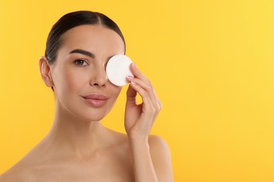 Beautiful woman removing makeup with cotton pad on yellow background, space for text