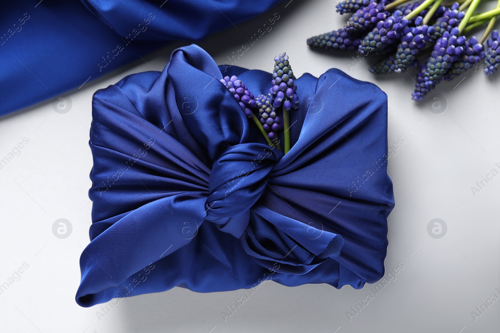 Photo of Furoshiki technique. Gift packed in blue silk fabric, muscari flowers and ribbon on white table, flat lay