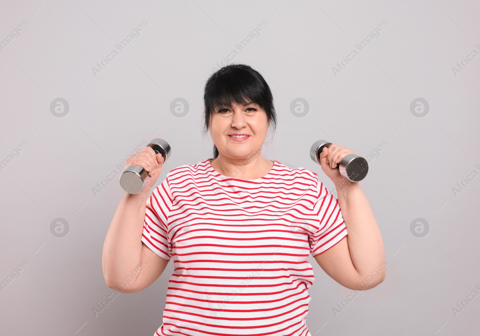 Photo of Happy overweight mature woman doing exercise with dumbbells on grey background