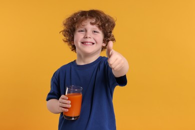 Photo of Cute little boy with glass of fresh juice showing thumb up on orange background