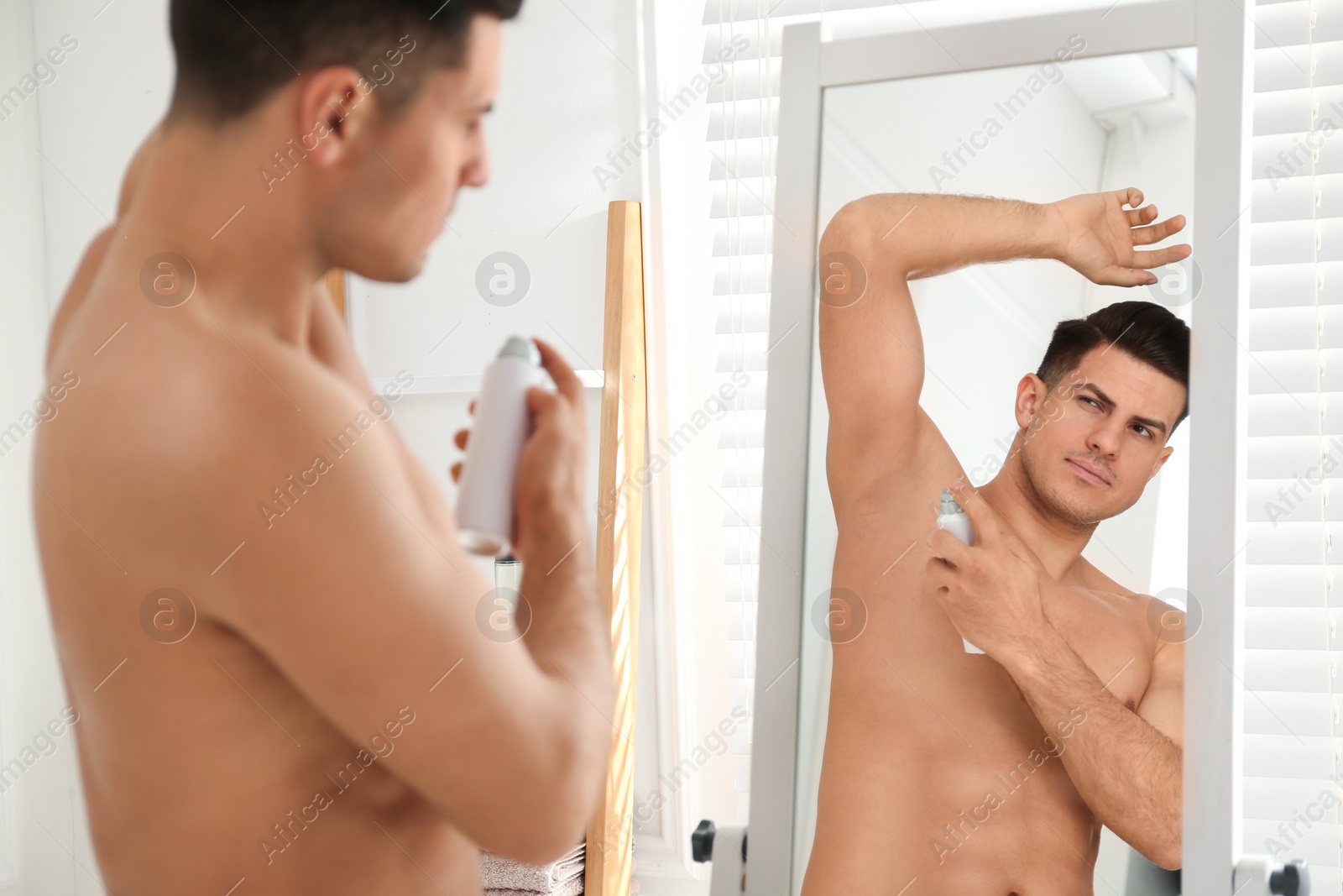 Photo of Handsome man applying deodorant to armpit near mirror at home