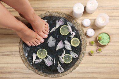 Woman soaking her feet in plate with water, flower petals and lime slices on wooden floor, top view. Pedicure procedure