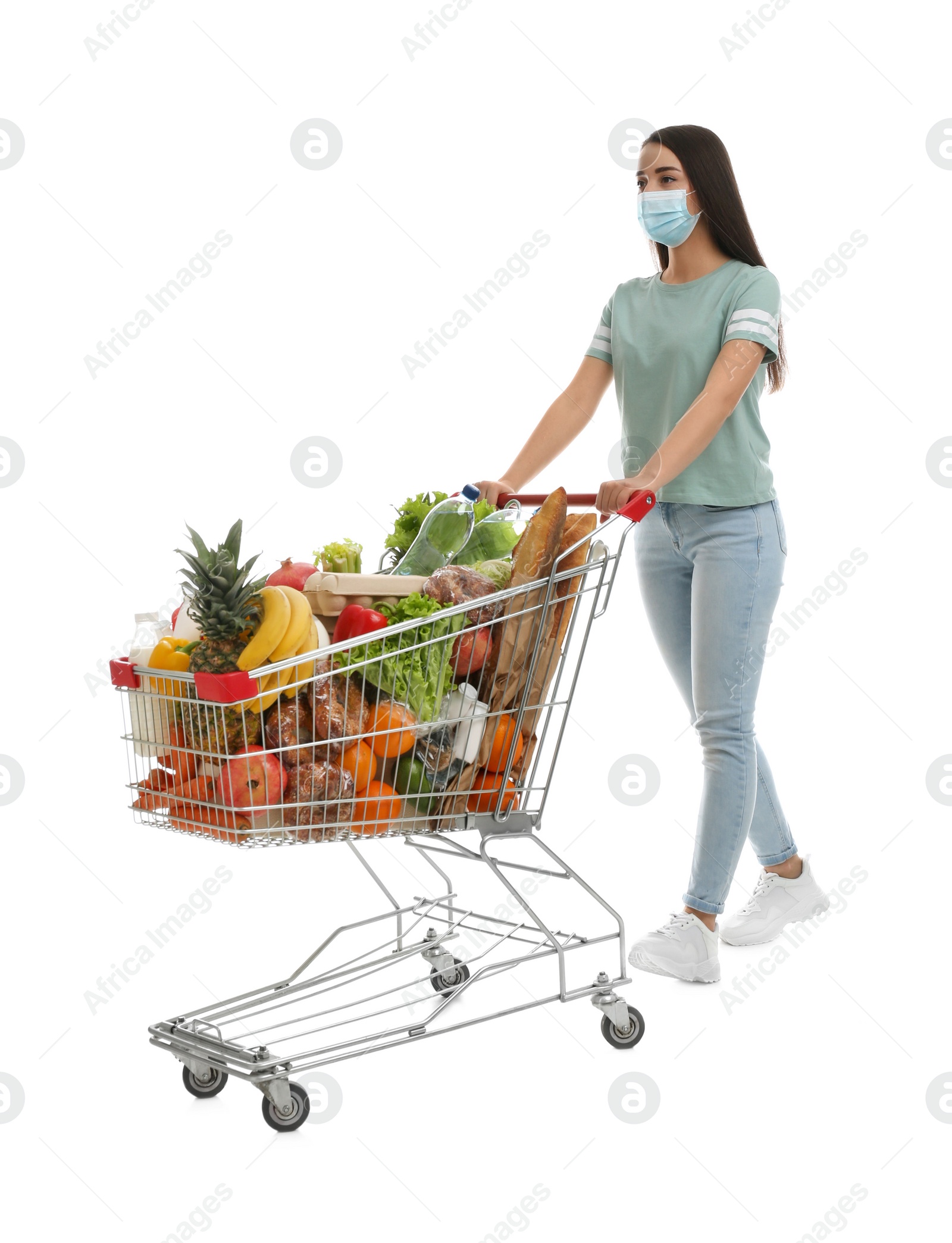 Photo of Young woman in medical mask with shopping cart full of groceries on white background