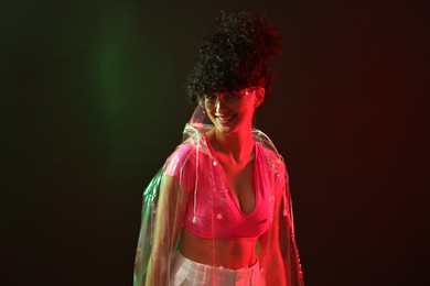 Photo of Beautiful young woman in transparent coat and sunglasses posing on color background in neon lights