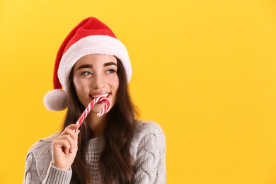 Beautiful woman in Santa Claus hat holding candy cane on yellow background. Space for text