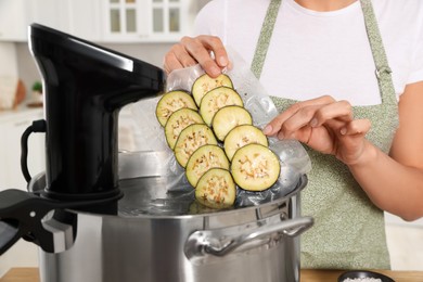 Photo of Woman putting vacuum packed eggplants into pot with sous vide cooker in kitchen, closeup. Thermal immersion circulator