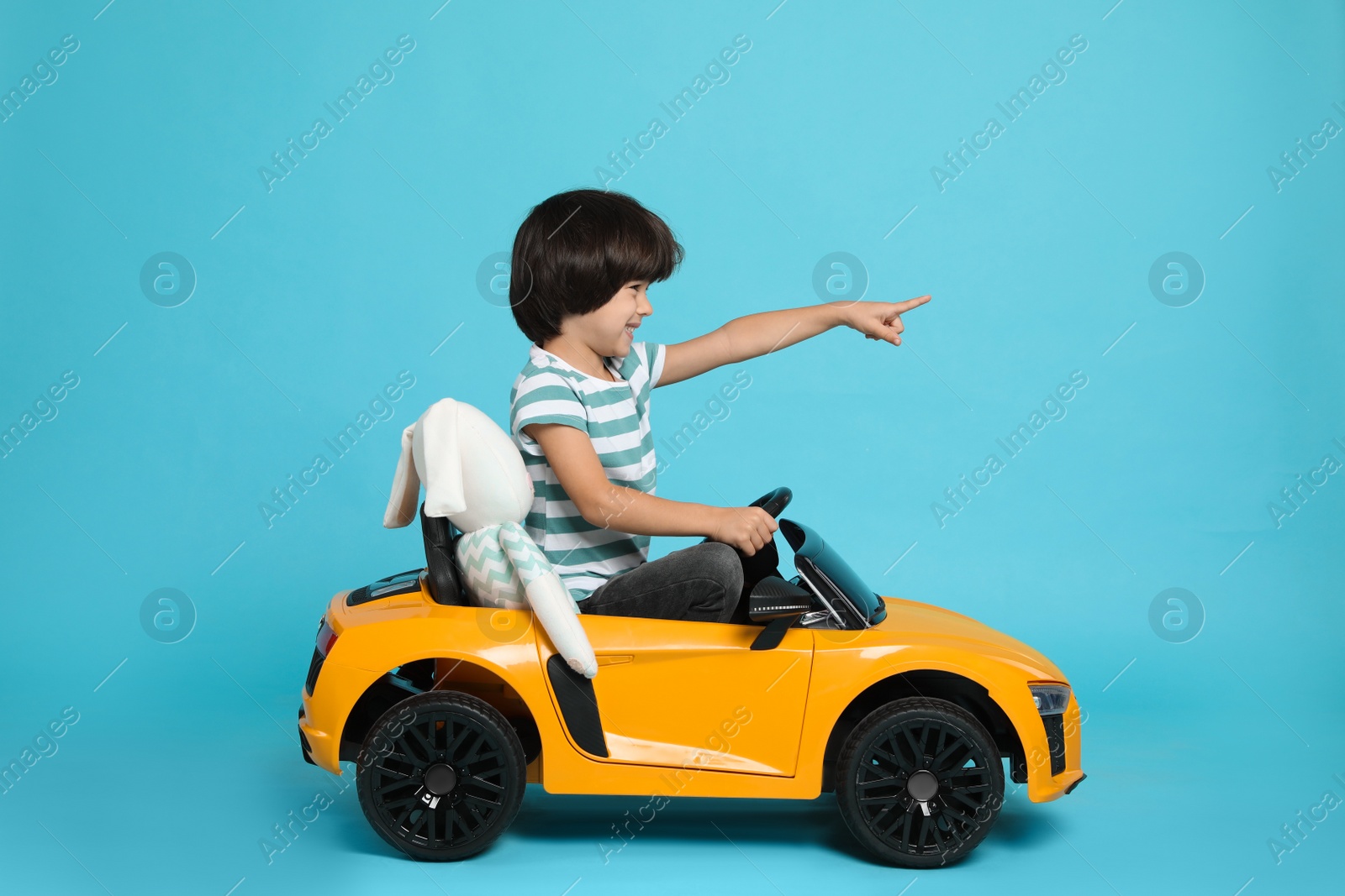 Photo of Cute little boy with toy bunny driving children's car on light blue background