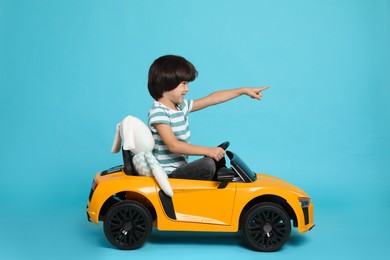 Cute little boy with toy bunny driving children's car on light blue background