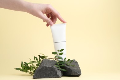 Photo of Woman with tube of cream, branches and stones on light yellow background, closeup