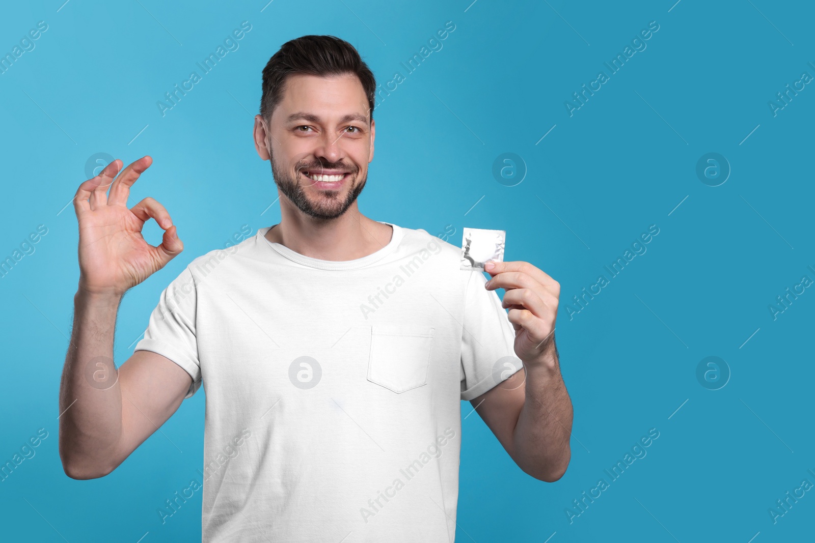 Photo of Happy man with condom showing ok gesture on light blue background. Space for text