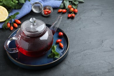 Photo of Teapot with aromatic rose hip tea, berries and mint on black table, space for text