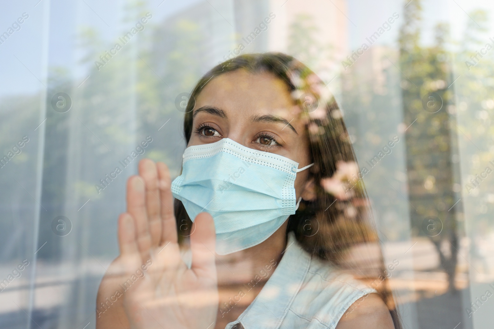 Photo of Stressed woman in protective mask looking out of window, view through glass. Self-isolation during COVID-19 pandemic