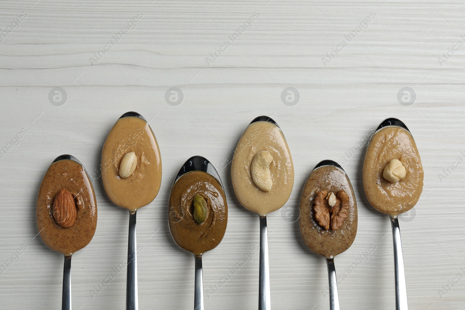 Photo of Tasty nut butters in spoons and raw nuts on white wooden table, flat lay. Space for text