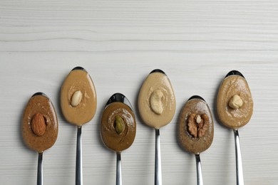 Tasty nut butters in spoons and raw nuts on white wooden table, flat lay. Space for text