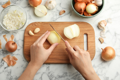 Photo of Woman cutting fresh onion on wooden board at white marble table, top view