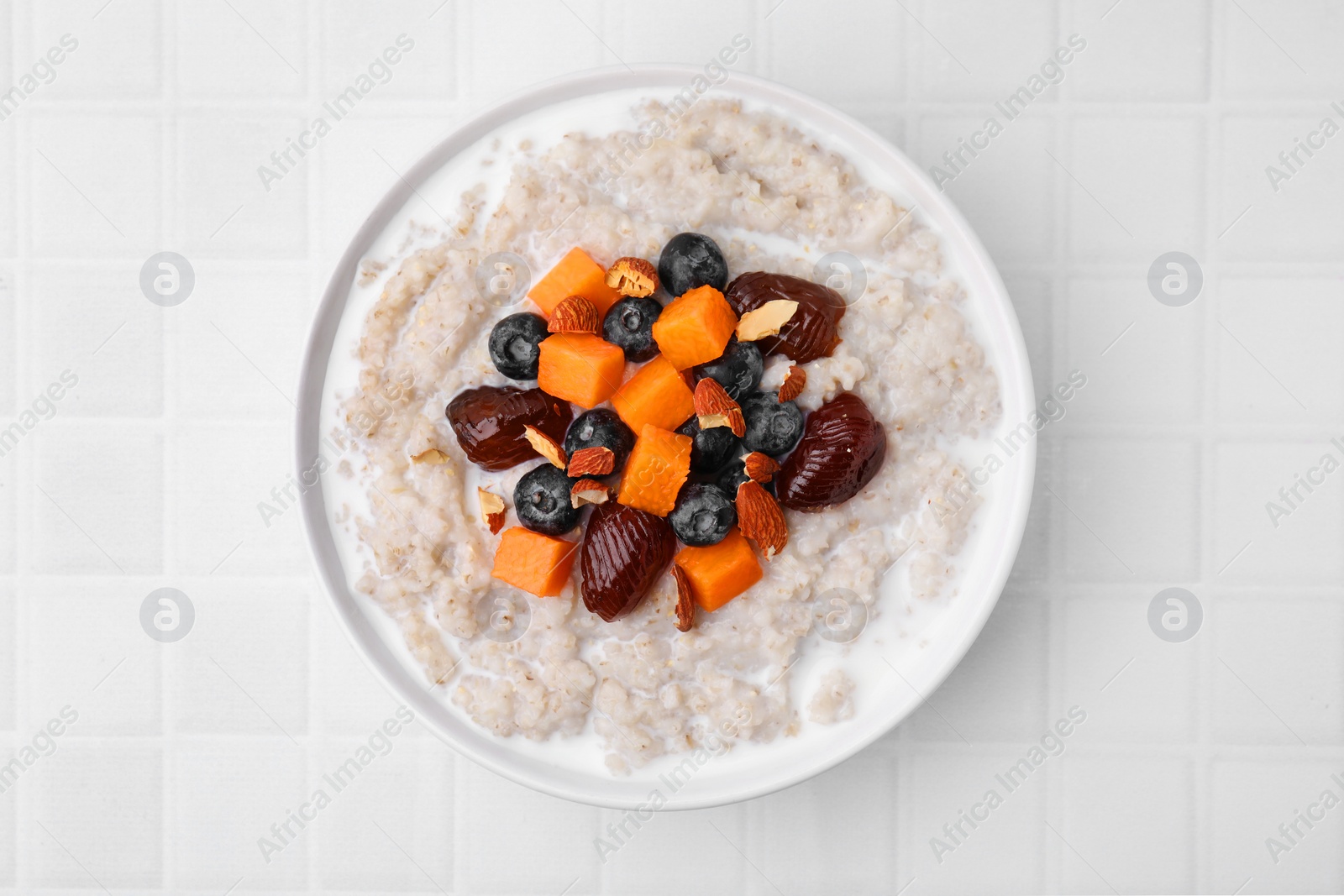 Photo of Delicious barley porridge with blueberries, pumpkin, dates and almonds in bowl on white tiled table, top view