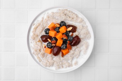 Photo of Delicious barley porridge with blueberries, pumpkin, dates and almonds in bowl on white tiled table, top view