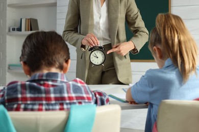 Photo of Teacher scolding pupils for being late in classroom, focus on alarm clock