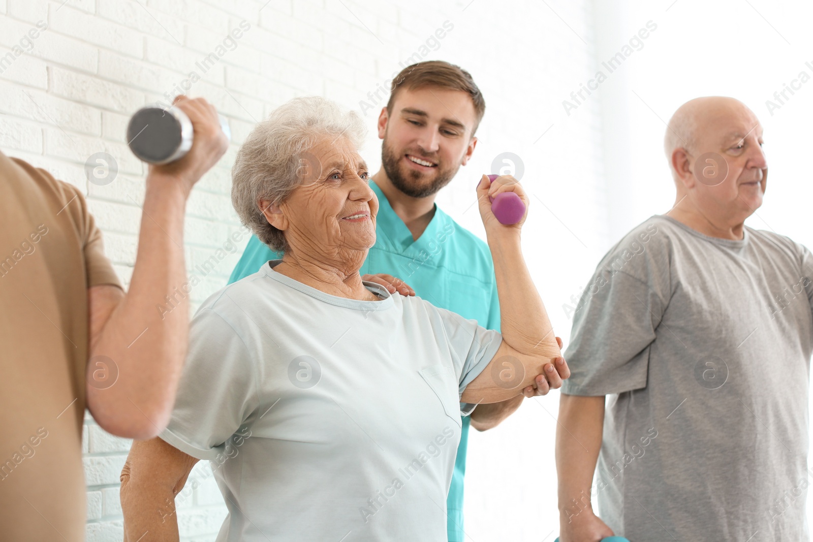 Photo of Care worker helping elderly woman to do exercise with dumbbell in hospital gym