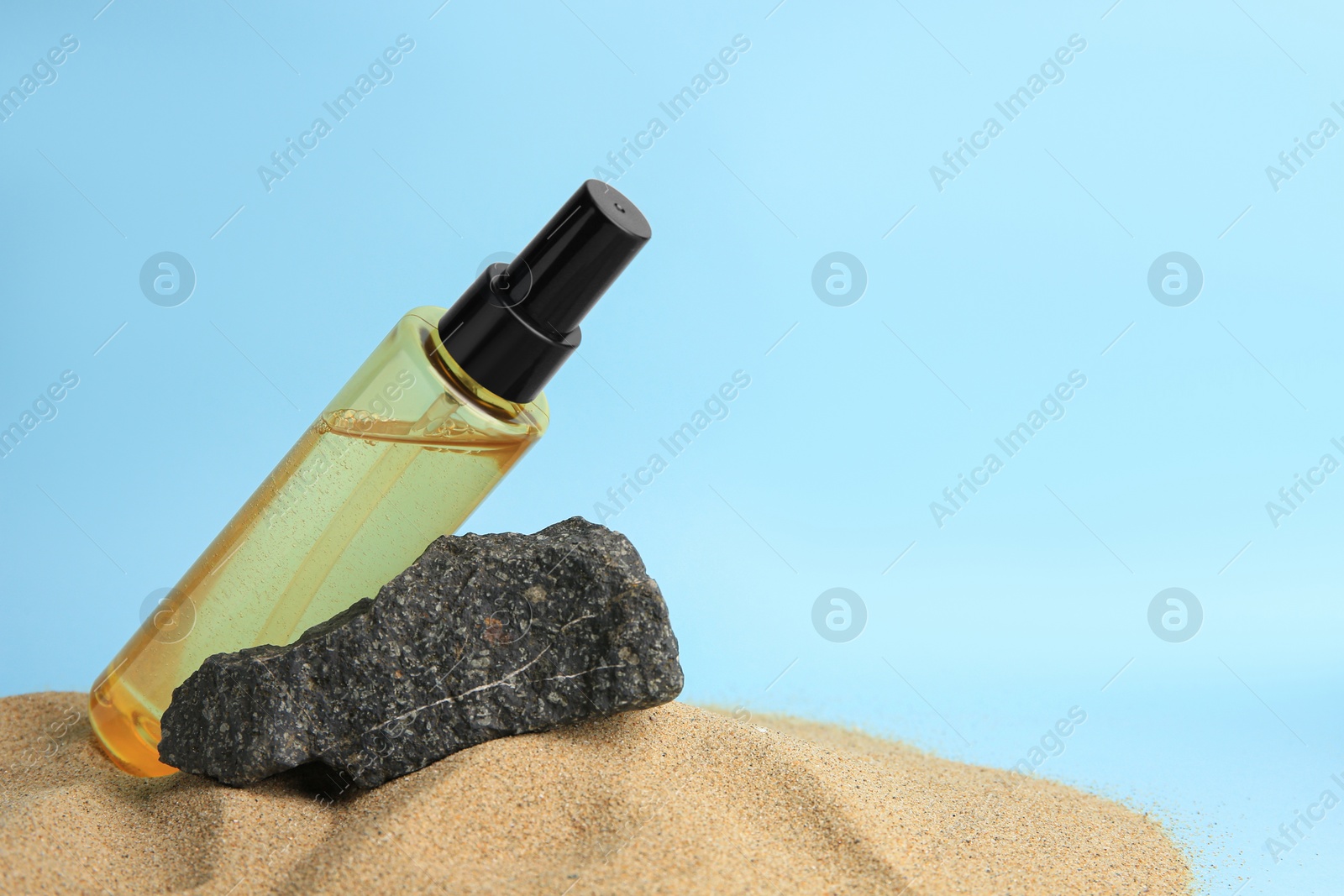 Photo of Bottle with serum and stone on sand against light blue background, space for text. Cosmetic product