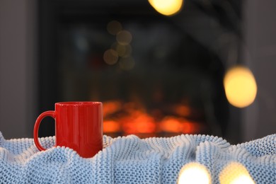 Cup of hot drink on knitted blanket near fireplace at home, space for text. Cozy atmosphere