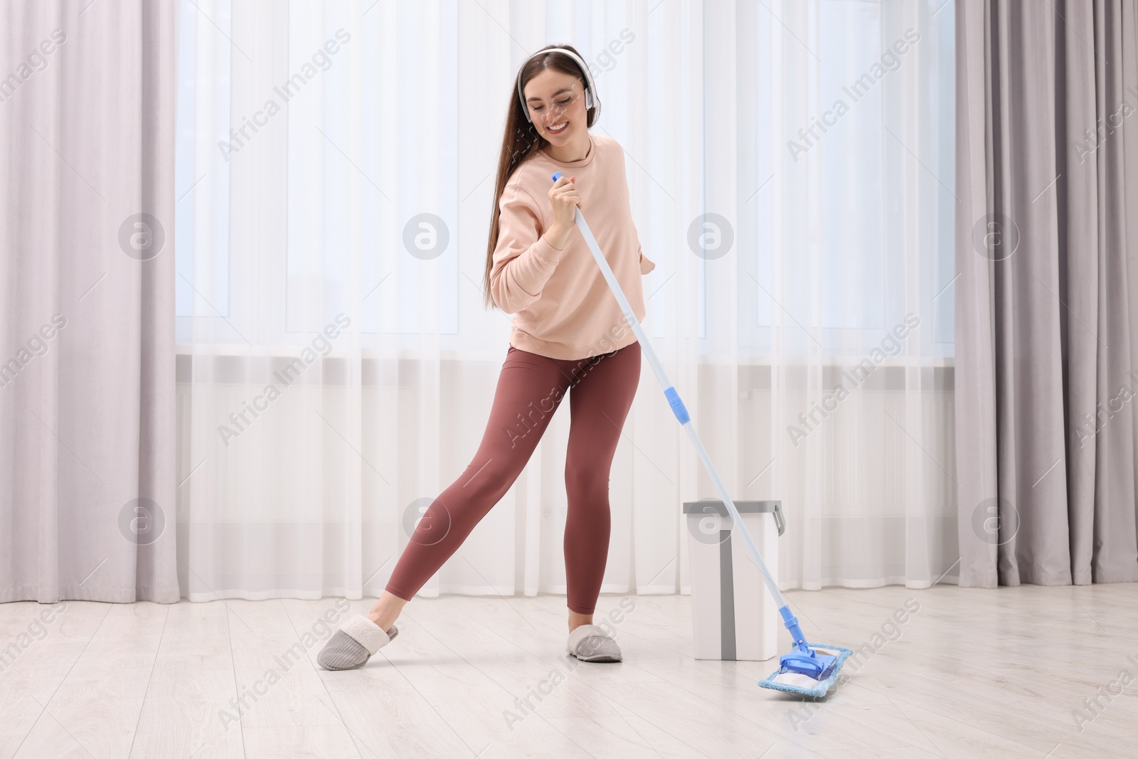 Photo of Enjoying cleaning. Happy woman in headphones listening music and mopping indoors