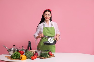 Photo of Young housewife with pan, spatula and products on pink background