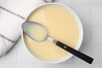Photo of Bowl with condensed milk and spoon on white tiled table, top view