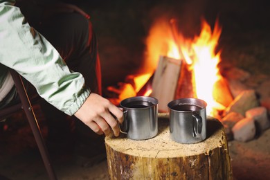 Photo of Woman taking cup of hot drink near bonfire outdoors, closeup. Camping season