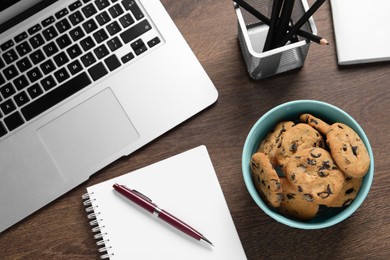 Bowl with chocolate chip cookies, laptop and office supplies on wooden table, flat lay